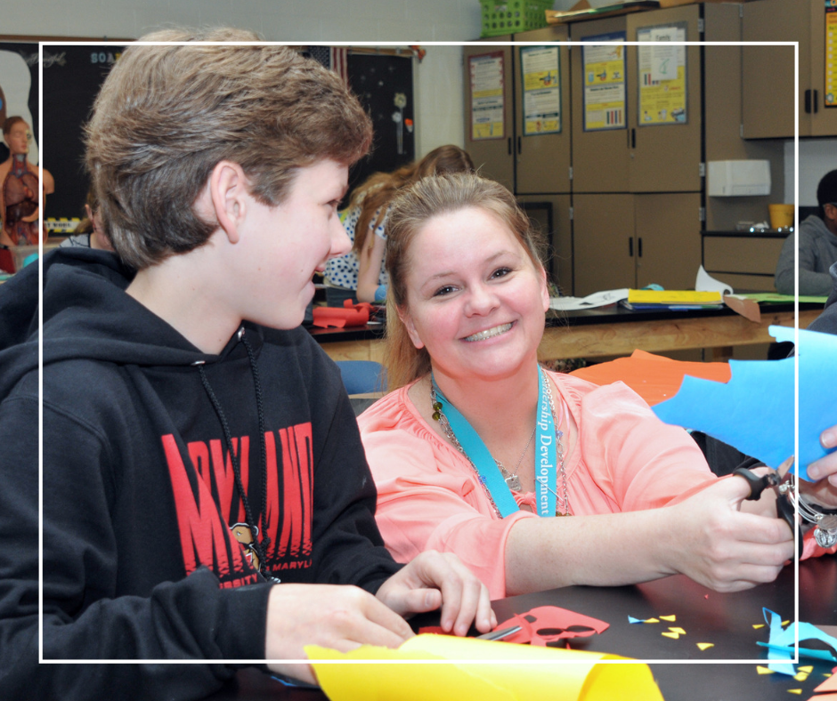 White, male student in middle school working with a white, female teacher on a craft project.
