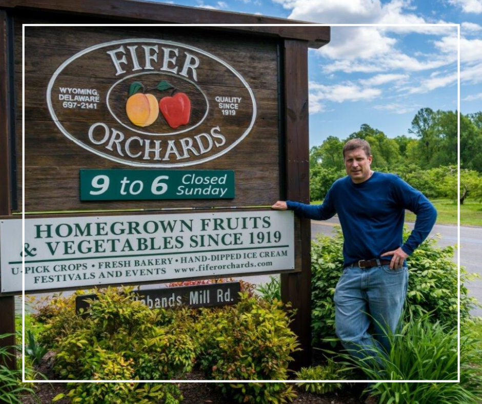 White, male business partner standing next to a sign that says Fifer Orchards, Homegrown Fruits and Vegetables since 1919