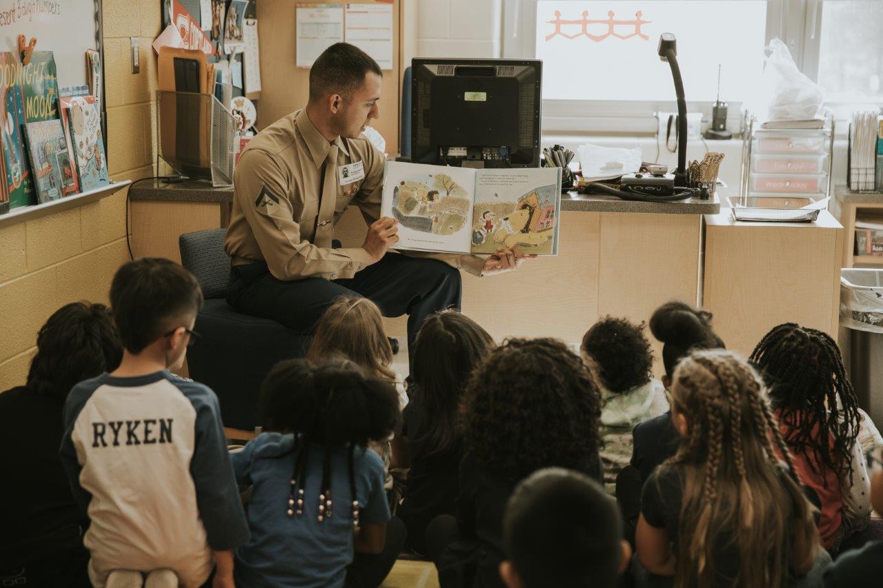 A male marine reads a picture book to a class of young students.