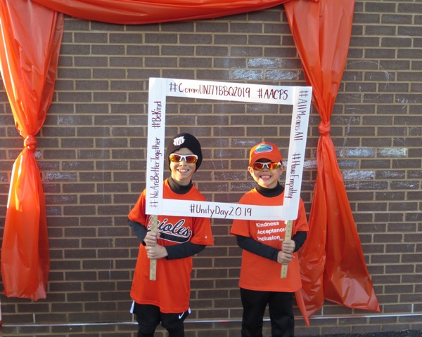 Two second grade boys stand holding a "CommUnityBBQ2019" photo frame under an orange curtain.